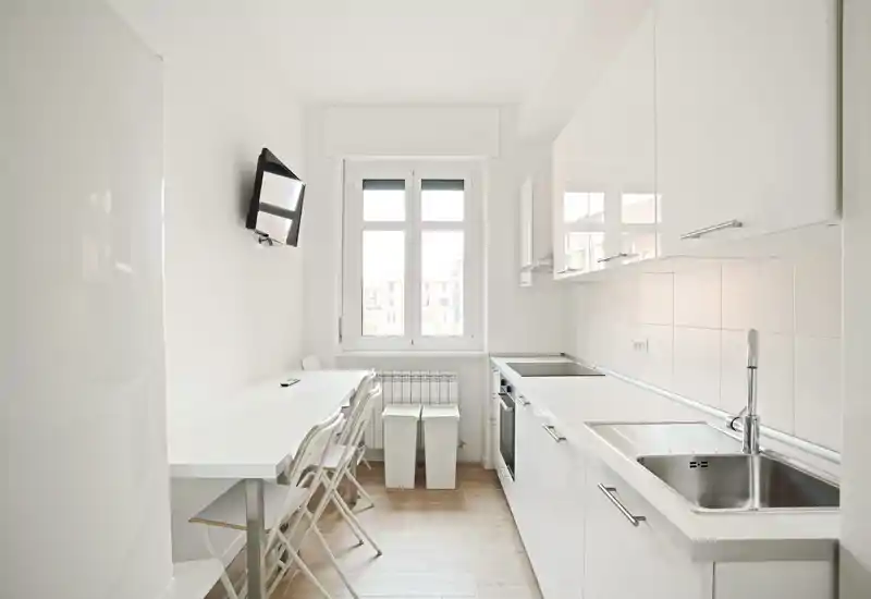 Cocina compacta y moderna, luminosa con armarios blancos, fregadero y horno integrados, y una pequeña mesa junto a la ventana — minimalista y perfecta para estancias cortas.