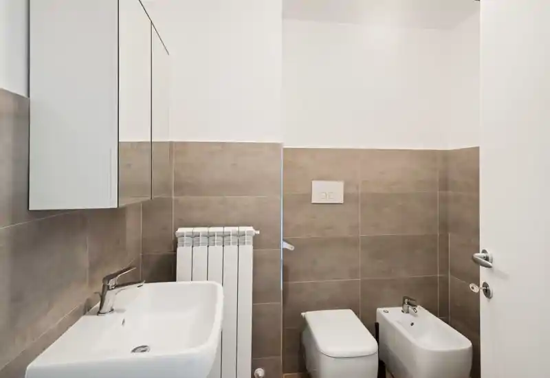 Baño moderno y limpio con lavabo, inodoro, bidé y armario con espejo; azulejos beige y sanitarios blancos que aportan un aspecto fresco y minimalista.