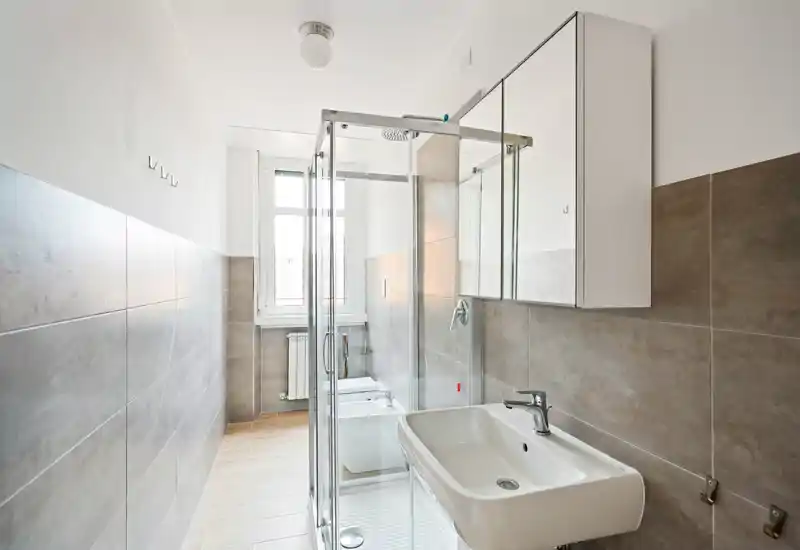 Baño moderno y luminoso con cabina de ducha de cristal, lavabo suspendido y armario con espejo. Baldosas neutras y una amplia ventana aportan luz natural y una estética limpia y contemporánea.