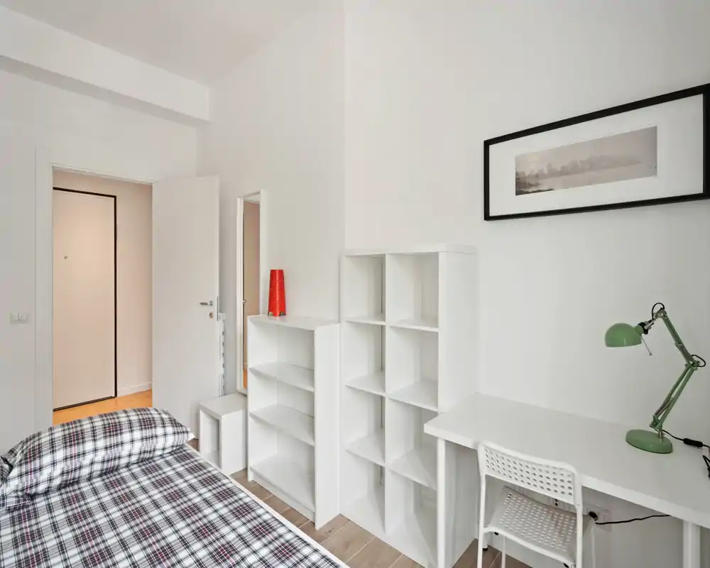 Camera da letto luminosa e minimal con un letto singolo, scaffalature aperte e una scrivania — ideale per uno studente o una persona sola. Pareti bianche e luce naturale donano un’atmosfera fresca e ariosa.
