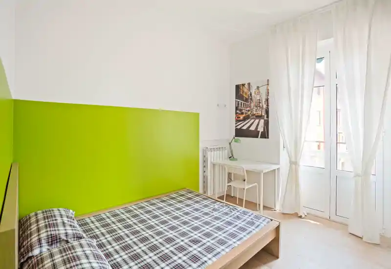 Camera da letto moderna e luminosa con letto matrimoniale davanti a una parete verde acceso, piccolo scrittoio e ampia finestra che illumina la stanza.