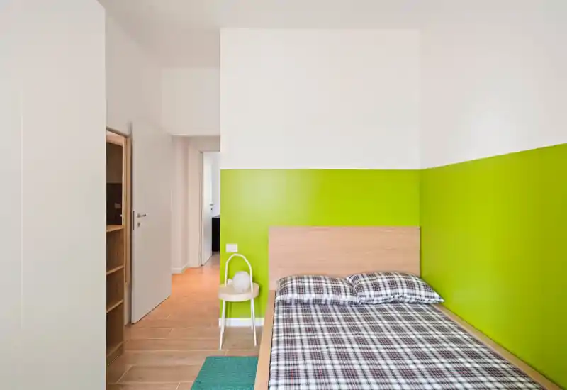 Camera da letto compatta e moderna con letto singolo in legno contro una parete verde acceso, biancheria a quadri e un piccolo sgabello come comodino; linee pulite e pavimento in legno caldo per un ambiente fresco e accogliente.
