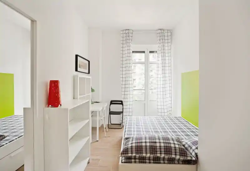 Camera da letto luminosa e minimale con letto singolo, copriletto a quadri, scrivania e scaffale vicino a una finestra alta che illumina la stanza con luce naturale.