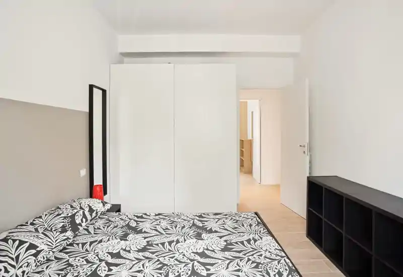 Dormitorio moderno y luminoso con cama doble estampada, armario blanco y puerta abierta al pasillo. Mobiliario minimalista y tonos neutros que aportan una atmósfera tranquila.