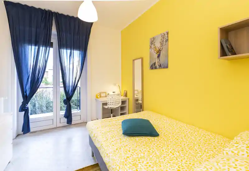 Sunny, well-staged bedroom with a double bed in the foreground, bright yellow accent wall, and a desk by the window letting in abundant natural light.