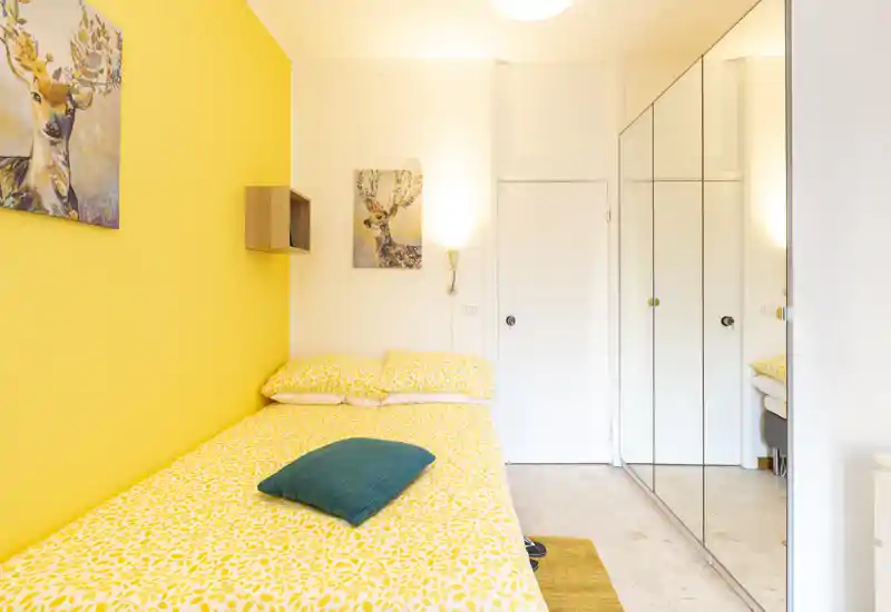 Bright, compact bedroom with a patterned yellow bedspread, matching pillows and a teal accent cushion. The mirrored wardrobe increases perceived space and the warm yellow wall and artwork create a cheerful, cozy atmosphere.