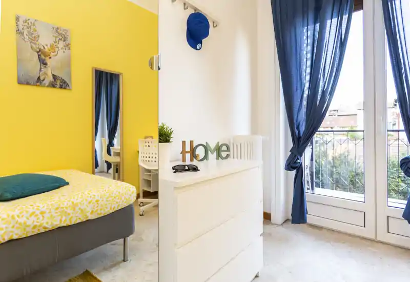 Bright, cheerful bedroom with a patterned bed, teal pillow and sunny yellow accent wall. Large balcony doors with navy curtains flood the room with natural light; clean, modern furnishings create a welcoming space ideal for short-term rentals.