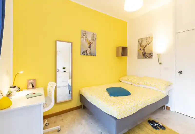 Sunny, well-staged bedroom with a double bed dressed in yellow patterned linens, a teal accent cushion, bedside wall lamp and a small desk — bright and welcoming.