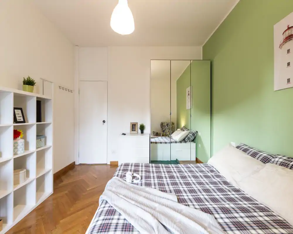 Camera da letto luminosa e ben arredata con letto matrimoniale, copriletto a quadri, armadio con ante a specchio e mensole — ambiente accogliente e arioso, ideale per affitti brevi.