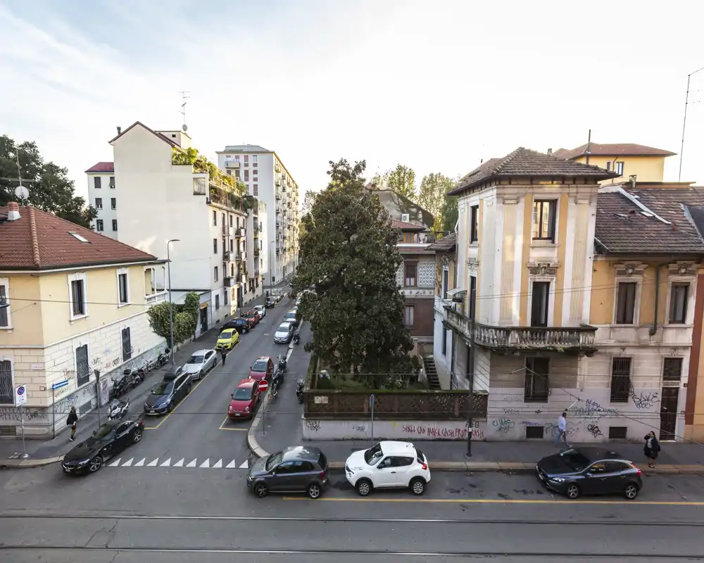 Vista urbana a livello strada con palazzine residenziali, auto parcheggiate e un grande albero; buona luce naturale e dettagli architettonici nitidi.