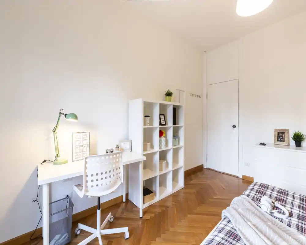Camera da letto luminosa e minimal con letto in primo piano, scrivania bianca e libreria a cubi per studio o deposito, pavimento in parquet a spina di pesce e illuminazione moderna.