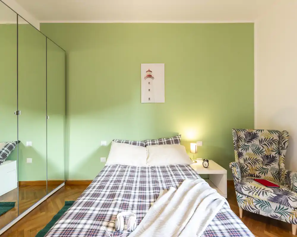 Camera da letto luminosa e accogliente con letto matrimoniale con copriletto a quadri, comodino con lampada e poltrona con fantasia. Armadio con ante a specchio e pavimento in parquet contribuiscono all'atmosfera calda.
