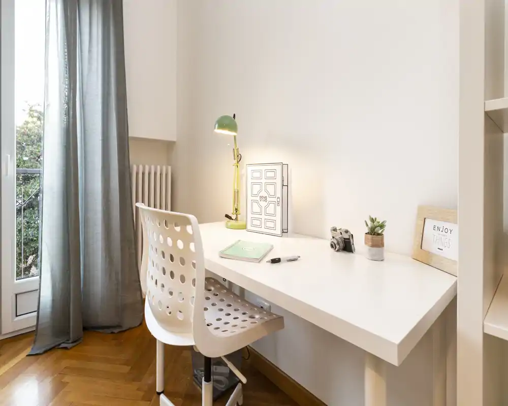 Spazio di lavoro luminoso e minimale con scrivania bianca, sedia traforata e lampada da tavolo vicino alla finestra. Piccoli dettagli decorativi come una pianta, una cornice con frase e una macchina fotografica vintage rendono l'angolo studio ordinato e funzionale.