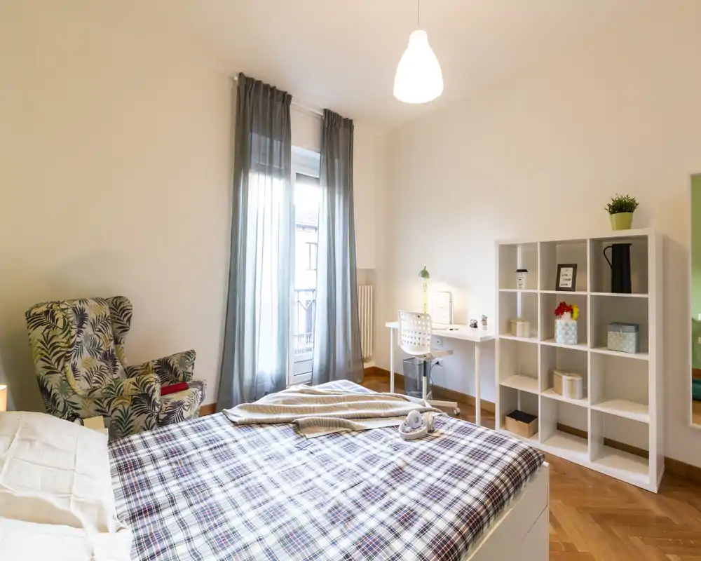 Camera da letto luminosa e ben arredata con letto ampio in primo piano, poltrona a fantasia, scrivania e scaffale: ideale per riposo e lavoro leggero.