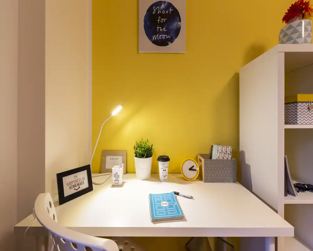 Angolo studio ordinato e ben illuminato con scrivania bianca, lampada da tavolo, pianta e contenitori decorativi su una parete gialla calda.