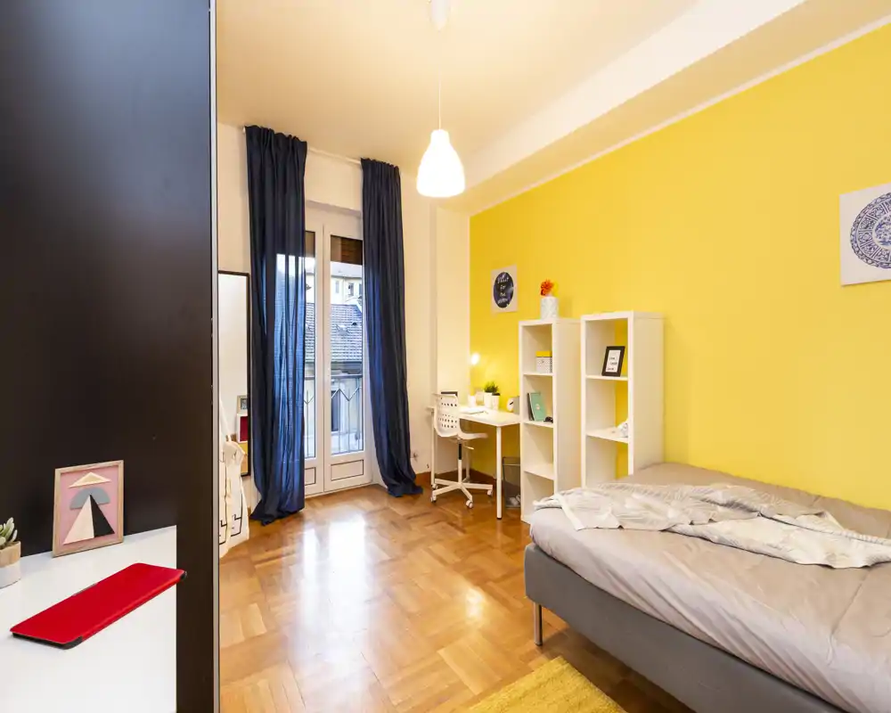Camera da letto luminosa e ben arredata con letto matrimoniale, parete gialla vivace, scaffalatura e una scrivania vicino alla finestra — ideale per viaggiatori singoli o coppie.