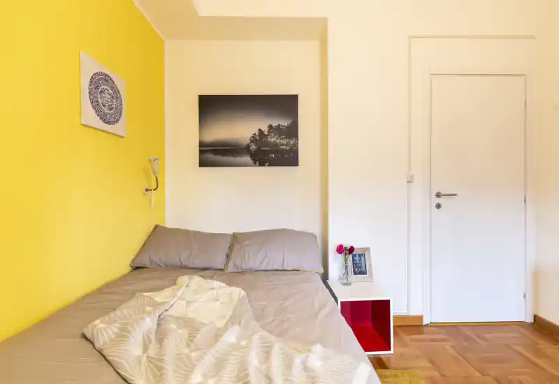 Bright, cozy bedroom with a double bed against a vivid yellow accent wall, bedside table with flowers and framed art — ideal for rental photos.