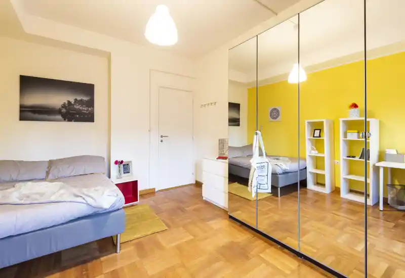 Sunny, well-styled bedroom with a comfortable double bed, warm yellow accent wall and large mirrored wardrobe that visually expands the space.