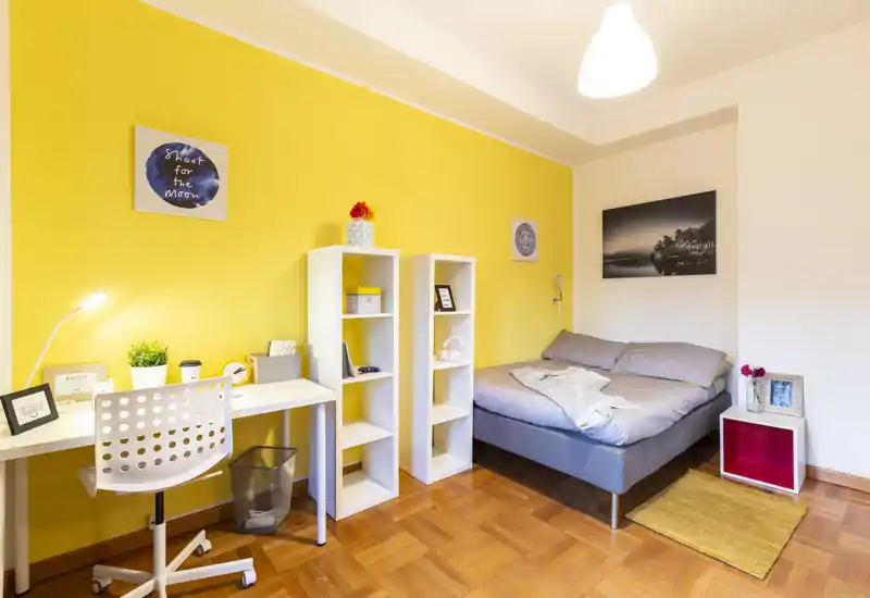Bright, cozy bedroom with a comfortable double bed, warm yellow accent wall, a small desk and shelving — ideal for a student or single professional.