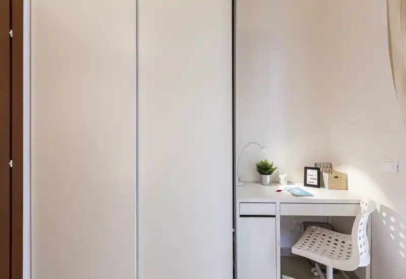 Angolo camera compatto con armadio a muro e piccolo scrittoio; stile minimale e ordinato con lampada da scrivania e pianta per un tocco accogliente.