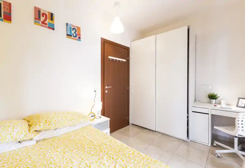 Camera da letto luminosa e ordinata con letto matrimoniale con biancheria gialla a fantasia, armadio bianco e una piccola scrivania — moderna e ben illuminata.