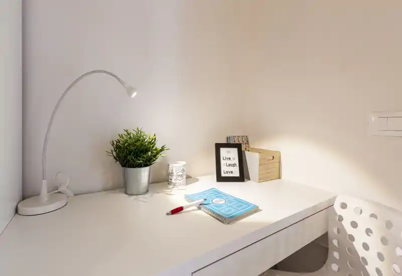 Angolo lavoro compatto e ben illuminato con scrivania bianca, lampada orientabile, piccolo vaso con pianta e cancelleria — un angolo ordinato ideale per studiare o lavorare da remoto.