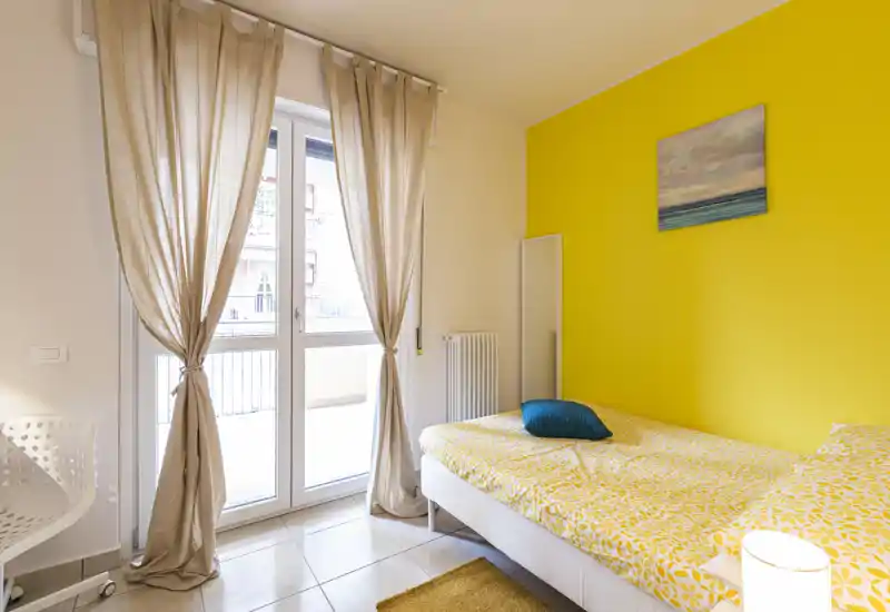 Camera da letto luminosa e moderna con parete gialla, letto singolo con biancheria fantasia, ampia finestra con tende leggere e piccolo tappeto — accogliente e ben illuminata.