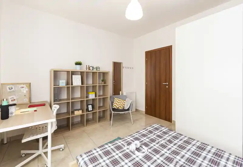 Camera da letto luminosa e ben arredata con copriletto a fantasia in primo piano, una scrivania per lo studio, una libreria a cubi e una poltroncina comoda — perfetta per una singola o uno studente.