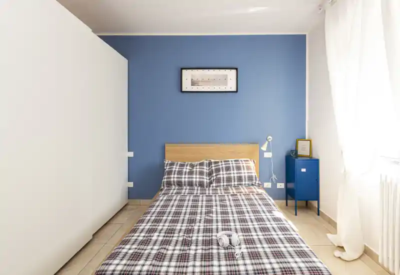 Camera da letto luminosa e moderna con letto matrimoniale centrato su una parete blu, biancheria a quadri, un comodino e luce naturale da una grande finestra.