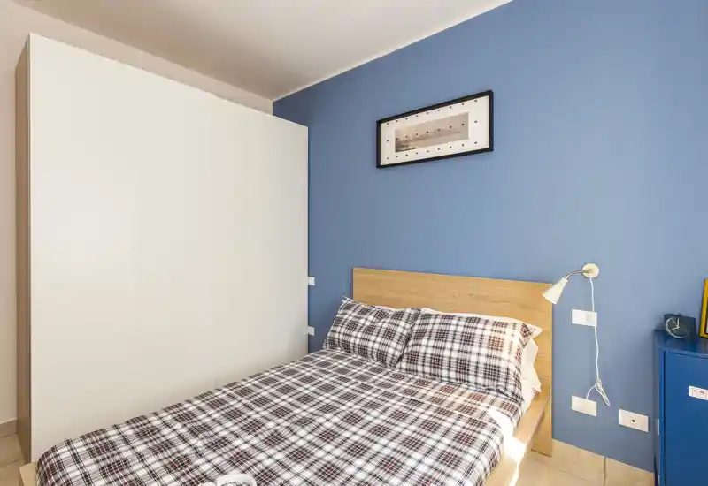 Camera da letto luminosa e ordinata con testiera in legno, copriletto a quadri, parete d'accento blu e un grande armadio bianco. Stile moderno e luce naturale che rendono la stanza accogliente.