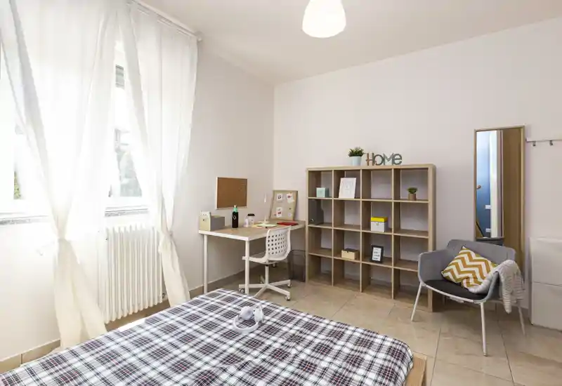 Camera da letto luminosa e ordinata con letto in primo piano, scrivania vicino alla finestra e scaffalatura a giorno per riporre; toni neutri e tessuti morbidi rendono l'ambiente accogliente.