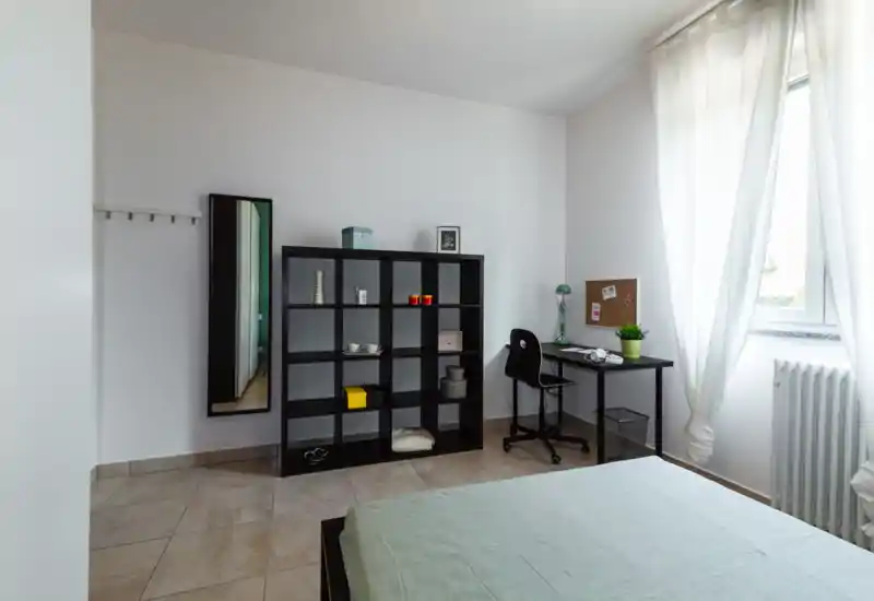 Dormitorio luminoso y minimalista con cama doble visible, estantería modular y un pequeño escritorio junto a la ventana con luz natural.