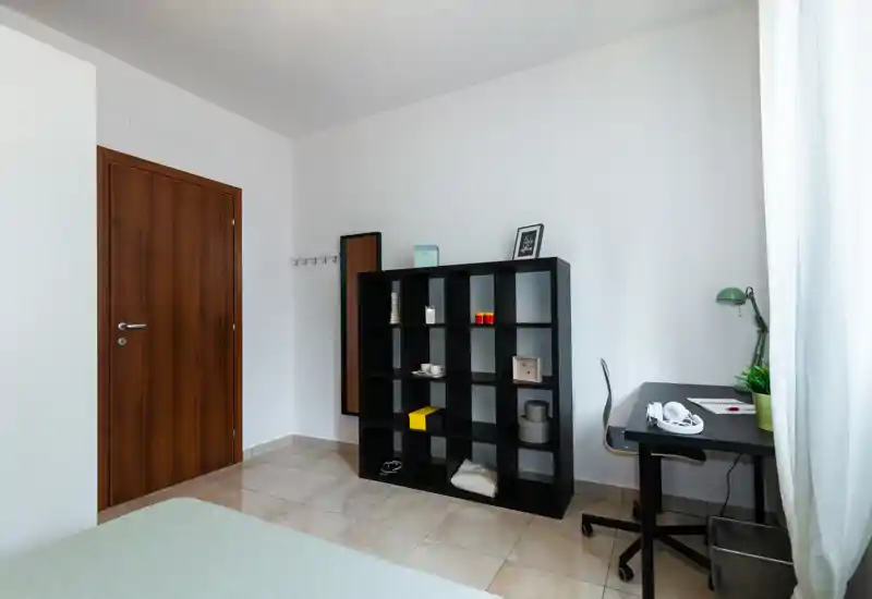Esquina de dormitorio luminosa y minimalista con el borde de la cama visible, una estantería negra y un escritorio junto a la ventana. Suelo de baldosas, paredes neutras y luz natural, apto para anuncios de alquileres de corta estancia.