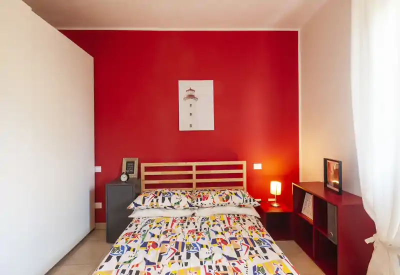 Camera da letto luminosa e ben arredata con letto matrimoniale al centro su una parete rossa d'accento, biancheria colorata, comodini e luce naturale morbida dalla finestra con tenda.