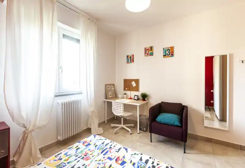 Camera da letto luminosa e ben allestita con copriletto colorato, una scrivania vicino alla finestra e una poltrona accogliente — perfetta per una persona o uno studente.