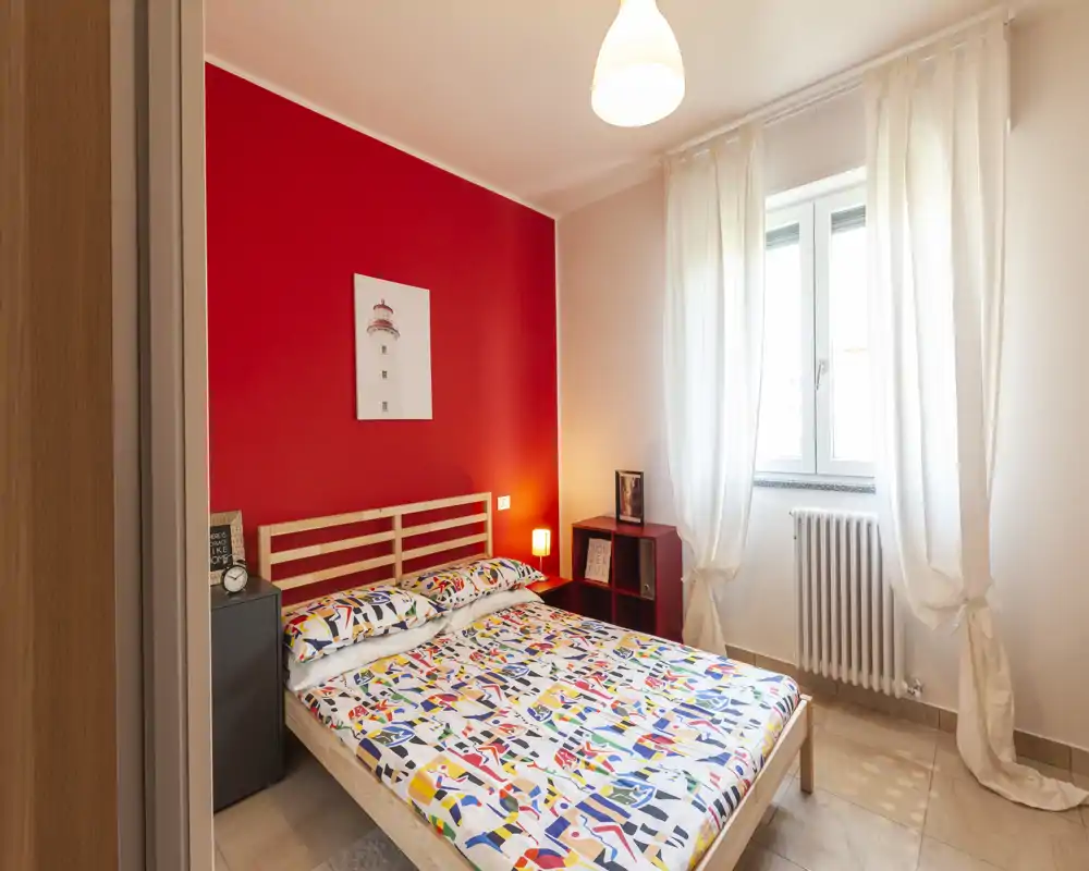 Camera da letto luminosa e ben arredata con letto matrimoniale contro una parete rossa a contrasto, biancheria dai motivi colorati, lampade da comodino e finestra con tende che garantisce luce naturale.