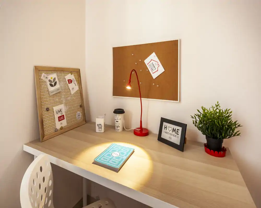 Angolo lavoro ordinato con scrivania in legno chiaro, lampada da tavolo rossa, bacheca in sughero e una piccola pianta: un punto luminoso e accogliente, ideale per studiare o lavorare da remoto.