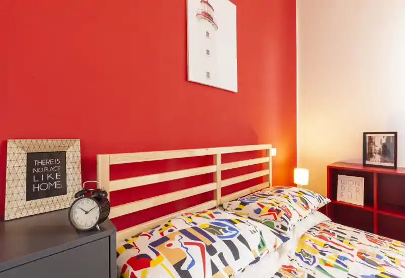 Camera da letto luminosa e colorata con testiera in legno a doghe, biancheria con fantasia e luce da comodino calda. La parete rossa e l'arredo curato creano un ambiente accogliente, perfetto per annunci.
