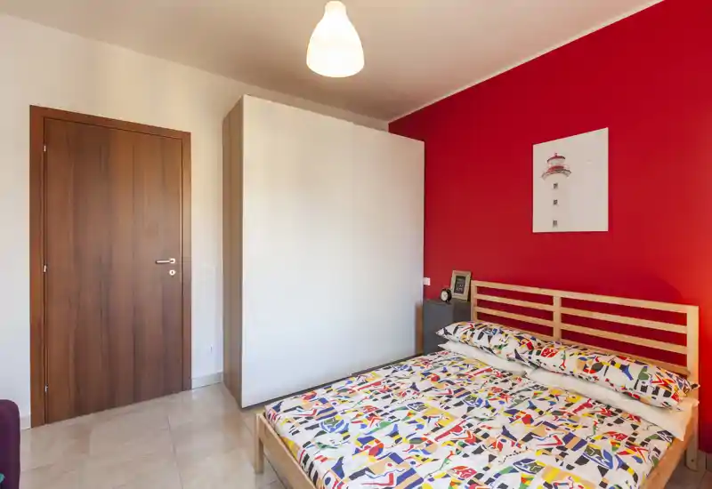 Camera da letto luminosa e moderna con struttura del letto in legno, biancheria colorata e un grande armadio bianco su una parete rossa.