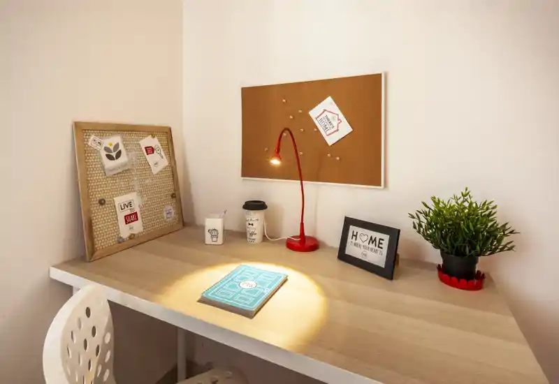 Angolo lavoro ordinato con scrivania in legno chiaro, lampada da tavolo rossa, bacheca in sughero e una piccola pianta: un punto luminoso e accogliente, ideale per studiare o lavorare da remoto.