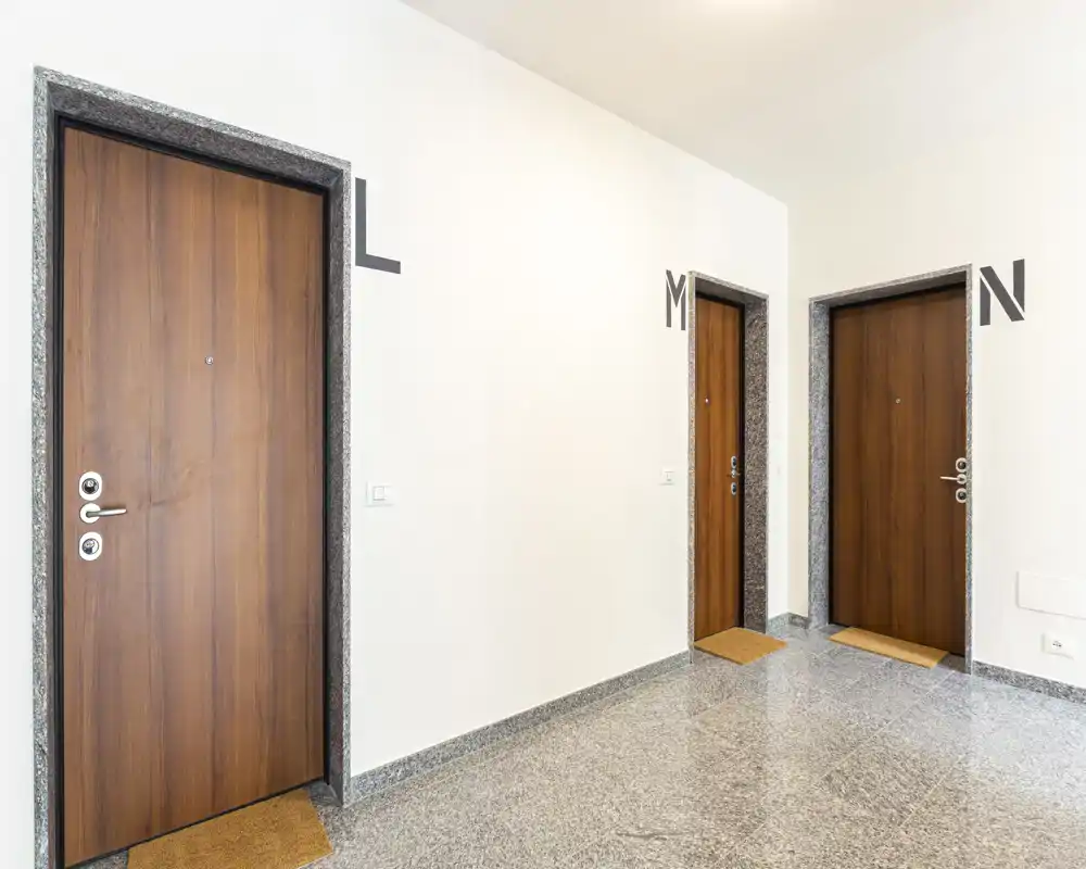 Ingresso/androne ben illuminato con tre porte in legno, pavimento in pietra pulito e pareti bianche neutre — pratico e di facile manutenzione.