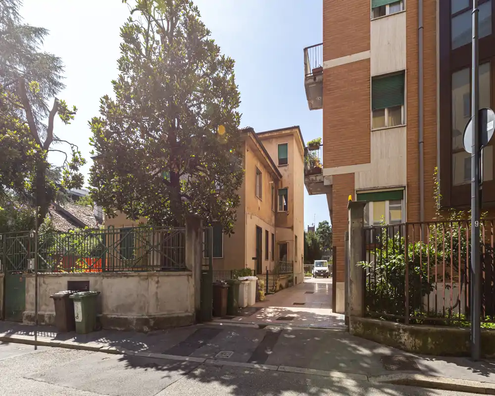 Ingresso/viottolo soleggiato tra edifici residenziali con accesso pedonale recintato, alberi e pavimentazione chiara — ideale per pubblicizzare un appartamento con ingresso privato.