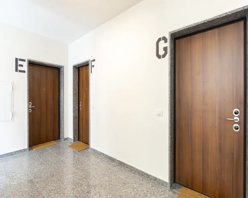 Corridoio d'ingresso luminoso e pulito con tre porte in legno etichettate E, F e G, pavimento in pietra levigata e pareti bianche — atrio condominiale ordinato.