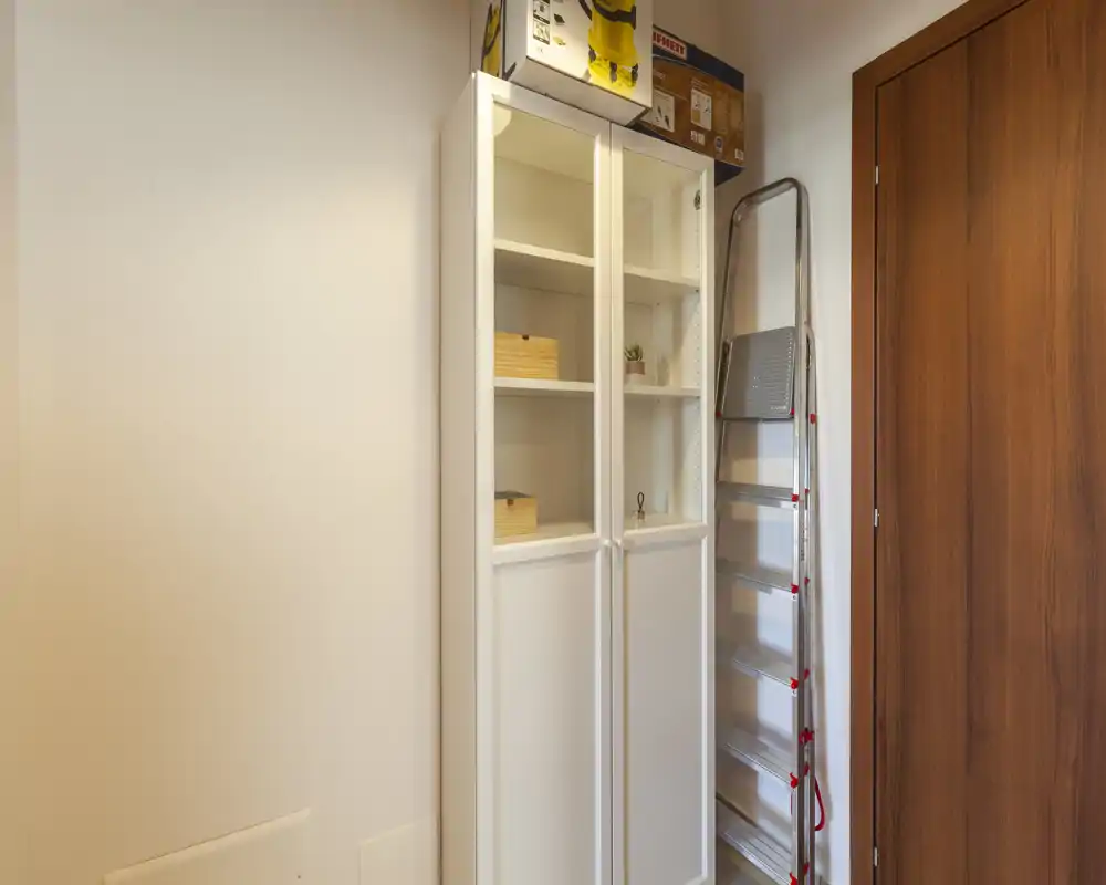 Piccolo angolo d'ingresso/corridoio con un armadietto alto bianco e una scala pieghevole accanto a una porta in legno; pareti neutre e arredo minimale.