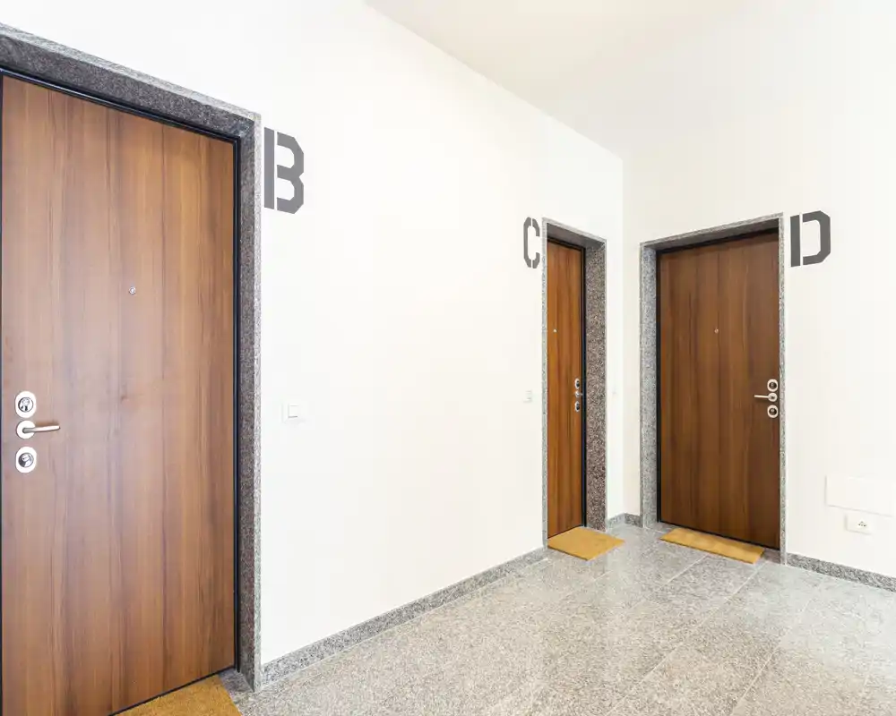 Corridoio d'ingresso comune luminoso con tre porte d'appartamento numerate in legno, pavimento in pietra e pareti bianche pulite — area di accesso ordinata e ben illuminata.
