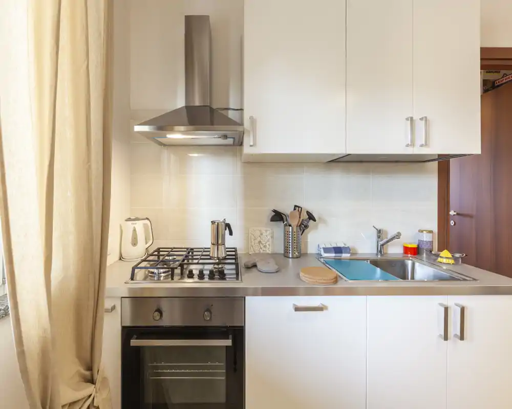 Cucina moderna compatta e luminosa con piano cottura a gas, cappa in acciaio, forno integrato e un lavello singolo — ben organizzata e pronta all'uso.