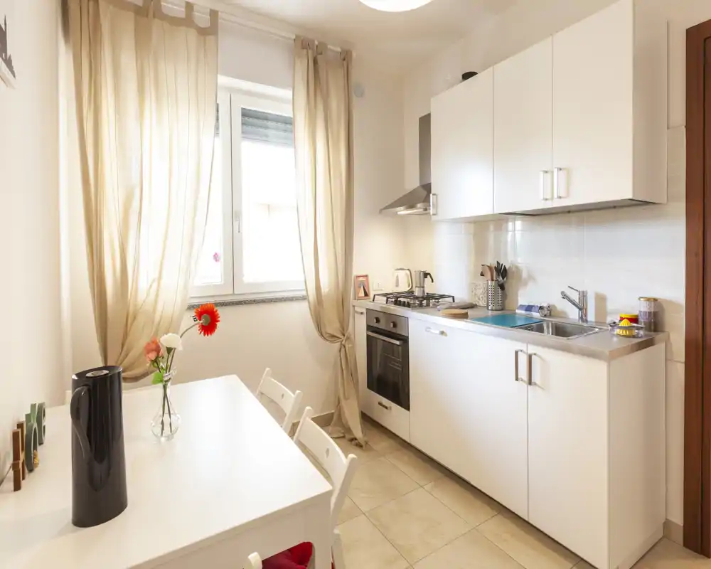 Cucina compatta e luminosa con mobili bianchi, forno incorporato e lavello, un piccolo tavolo da pranzo e luce naturale dalla finestra — ordinata e ben curata per un annuncio.