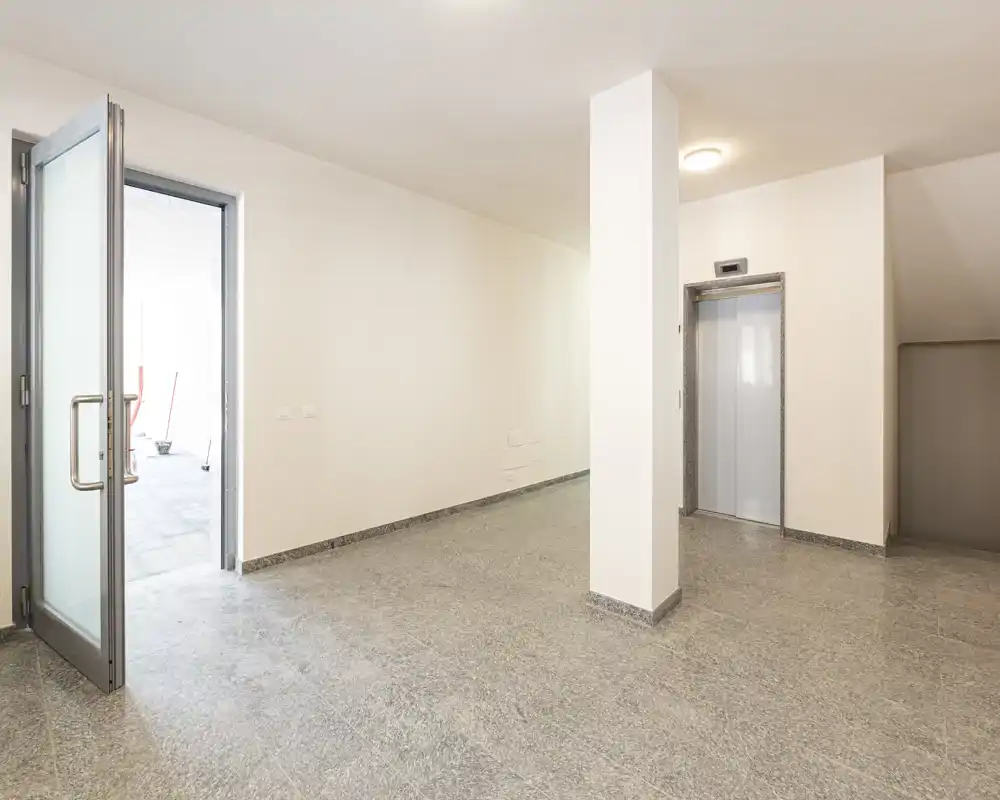 Ingresso/lobby moderno e luminoso con pavimento in piastrelle, ascensore e porta in vetro; pareti bianche neutre che creano un ambiente pulito e minimale adatto alle aree comuni.