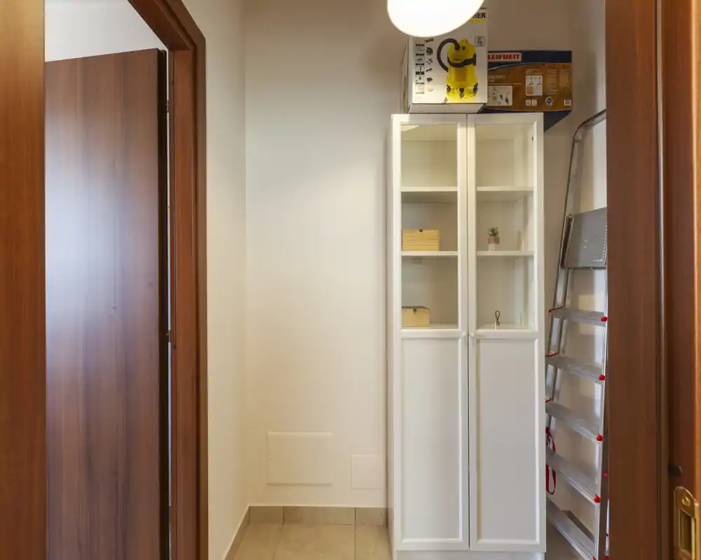 Piccolo ingresso con mobile contenitore bianco, pavimento in piastrelle e una scaletta richiudibile; ben illuminato e ordinato, adatto come anticamera o ripostiglio.