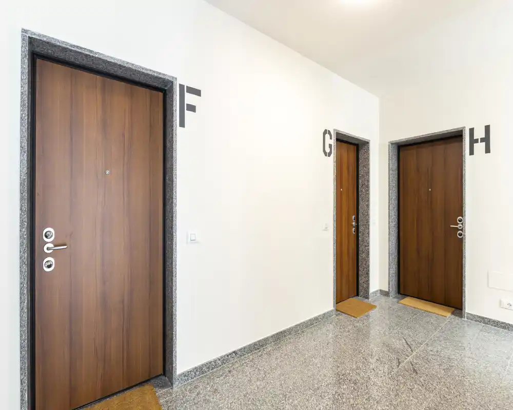 Corridoio d'ingresso condominiale ben illuminato con tre porte in legno (contrassegnate F, G, H), pavimento in pietra lucidato e pareti bianche pulite.